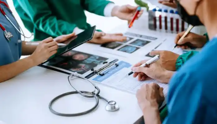 A collaborative medical team meeting representing Healthcare Industries by Grow Factor LLC, showing doctors and medical practitioners in blue and green scrubs gathered around a table reviewing brain scan MRIs, filling out clinical reports on a clipboard, using a tablet, and handling medical test tubes next to a stethoscope.