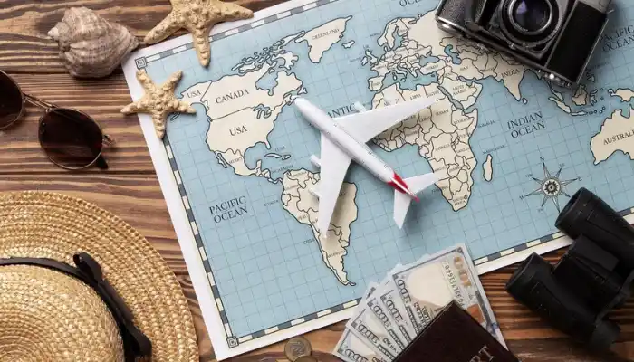A flat lay vacation planning layout representing Travel and Hospitality Industries by Grow Factor LLC, featuring a white toy airplane model resting in the center of a world map surrounded by travel gear including a straw hat, sunglasses, starfish, a vintage camera, binoculars, currency bills, and a passport on a wooden surface.